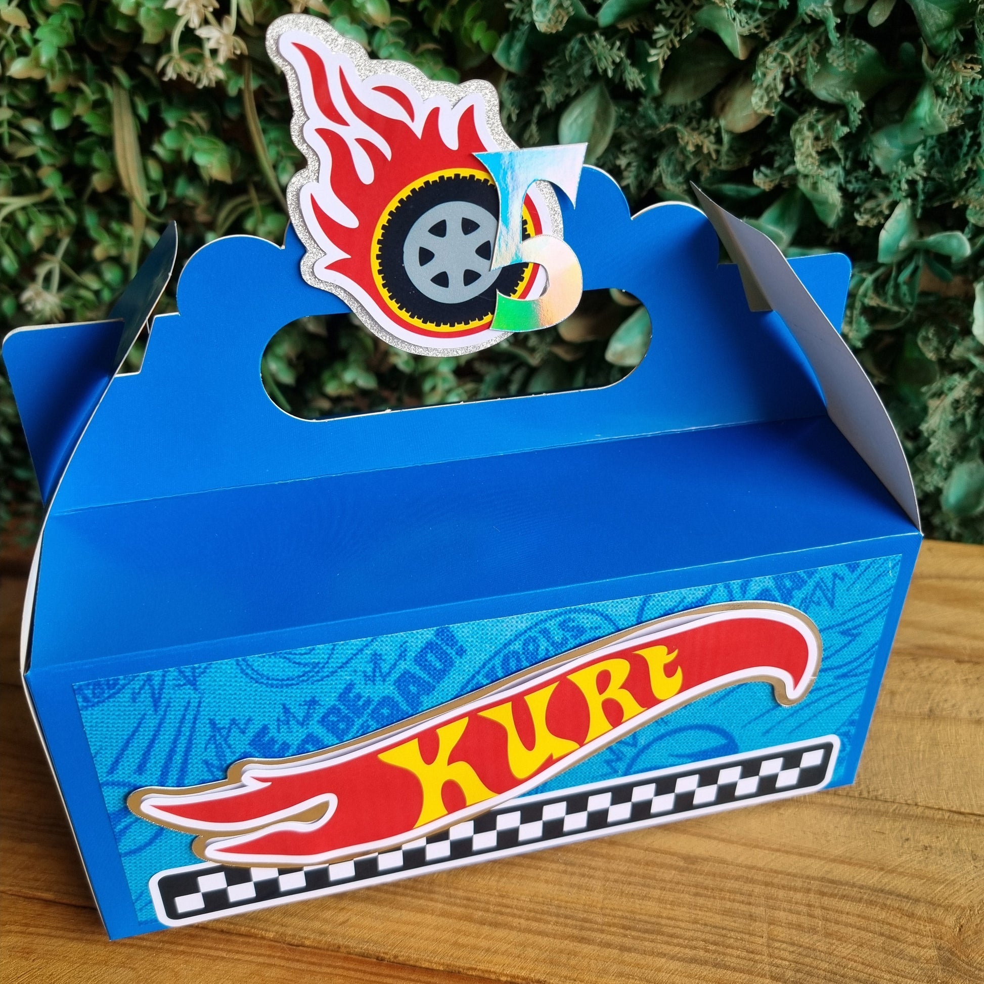 Blue toy car track with Hot Wheels logo on a wooden surface and green foliage background