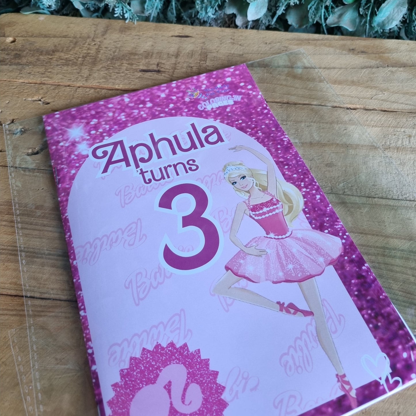 Pink birthday card with a girl in a dress and 'Alphula turns 3' text on a wooden surface.