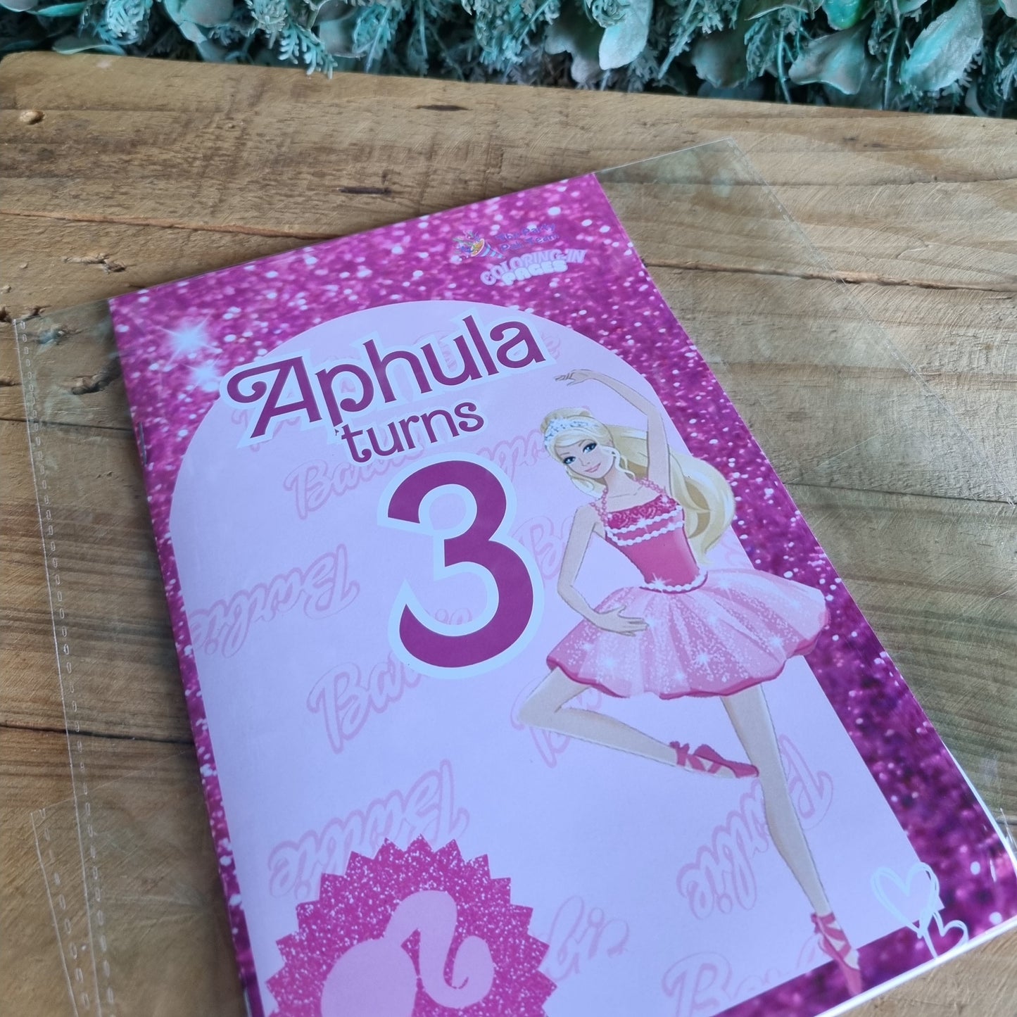 Pink birthday card with a doll design on a wooden surface next to green leaves.