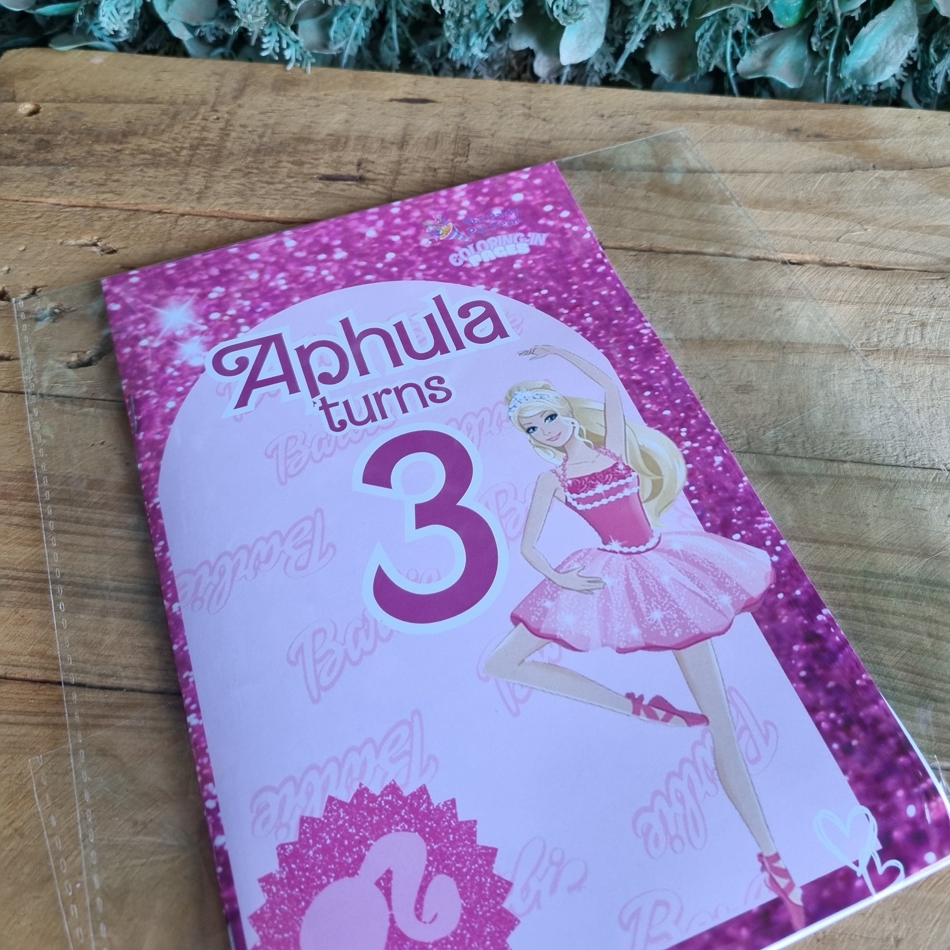 Pink birthday card with a doll design on a wooden surface next to green leaves.