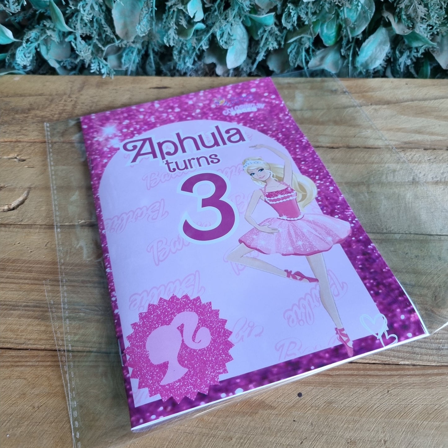 Pink birthday card with number 3 and fairy design on a wooden surface next to green leaves.