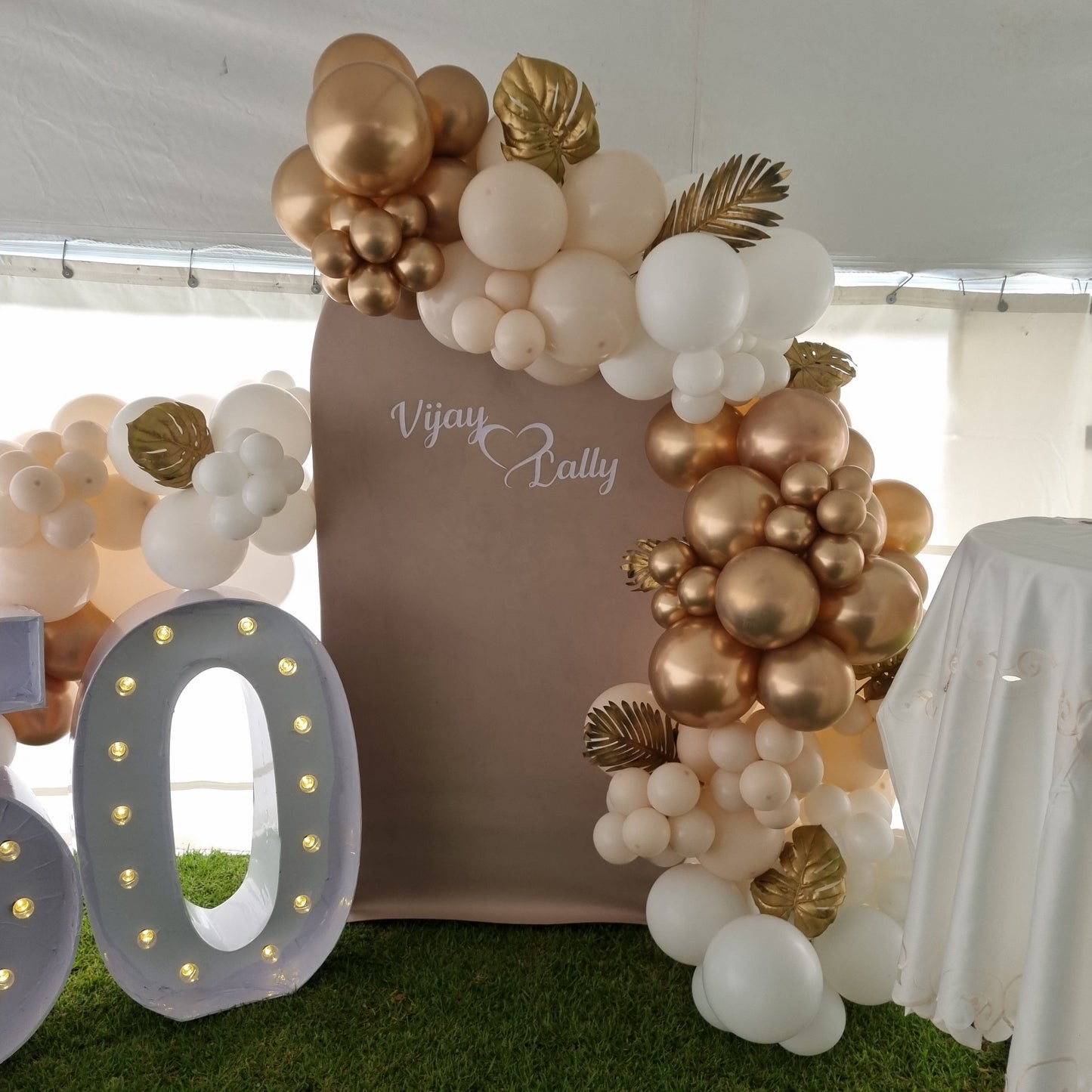 Decorative balloon arch with gold and white balloons and a number '0' light fixture.