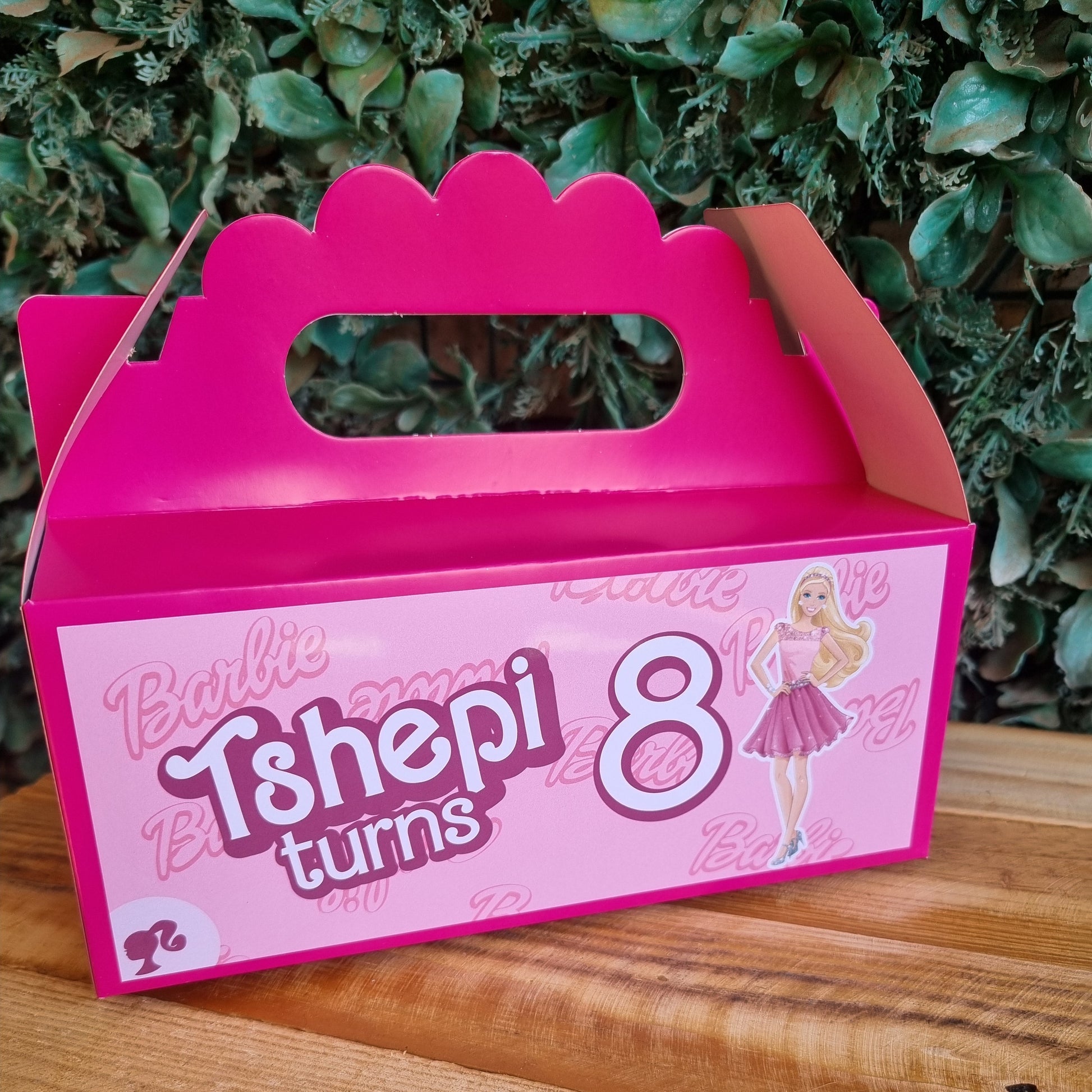 Pink birthday gift box with '8 turns 9' text on a wooden surface with greenery in the background