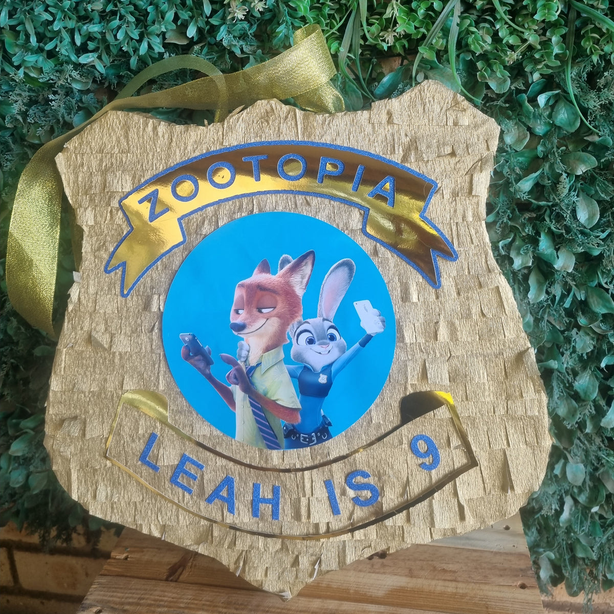 A custom wooden Zootopia piñata sign featuring Judy and Nick Wilde with the text 'ZOOTOPIA LEAH IS 9' and a gold ribbon hanging tag, set against a green foliage background.