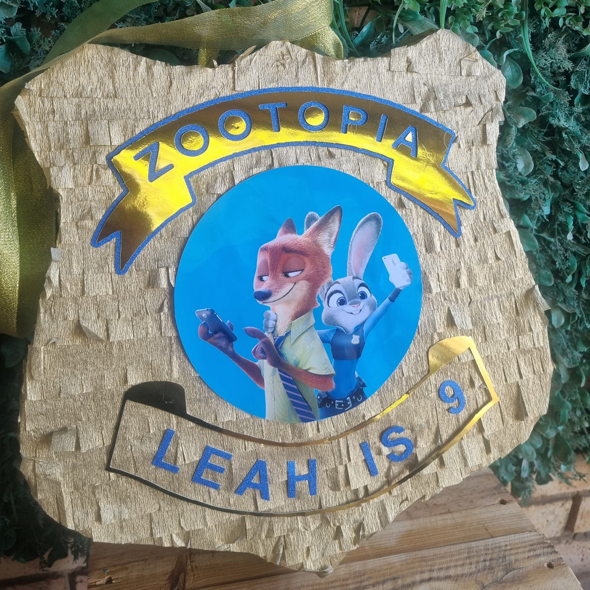 A custom Zootopia-themed wooden plaque or ornament featuring a cork-textured background, a circular blue insert with Judy Hopps and Nick Wilde, and gold lettering that reads 'ZOOTOPIA' and 'LEAH IS 9', hanging against green foliage.