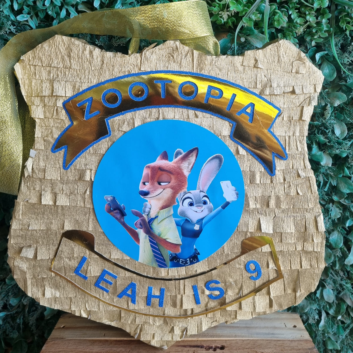 A custom Zootopia-themed wooden pinata shaped like a police badge, featuring the characters Judy Hopps and Nick Wilde on a blue circle, with 'ZOOTOPIA' and 'LEAH IS 9' in blue lettering. The pinata is decorated with a gold ribbon and hangs against a green hedge background.