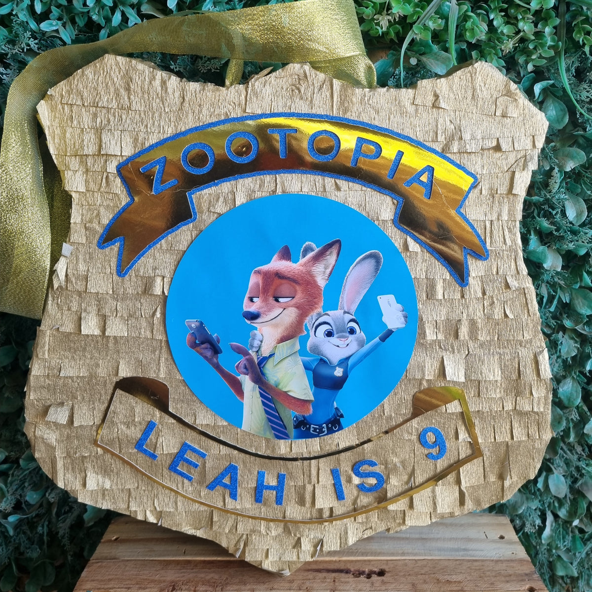 A custom Zootopia-themed wooden piñata shaped like the Zootopia Police Department badge, featuring a printed graphic of Judy Hopps and Nick Wilde with the text 'ZOOTOPIA' and 'LEAH IS 9', hanging on a wooden stand against a green foliage background.