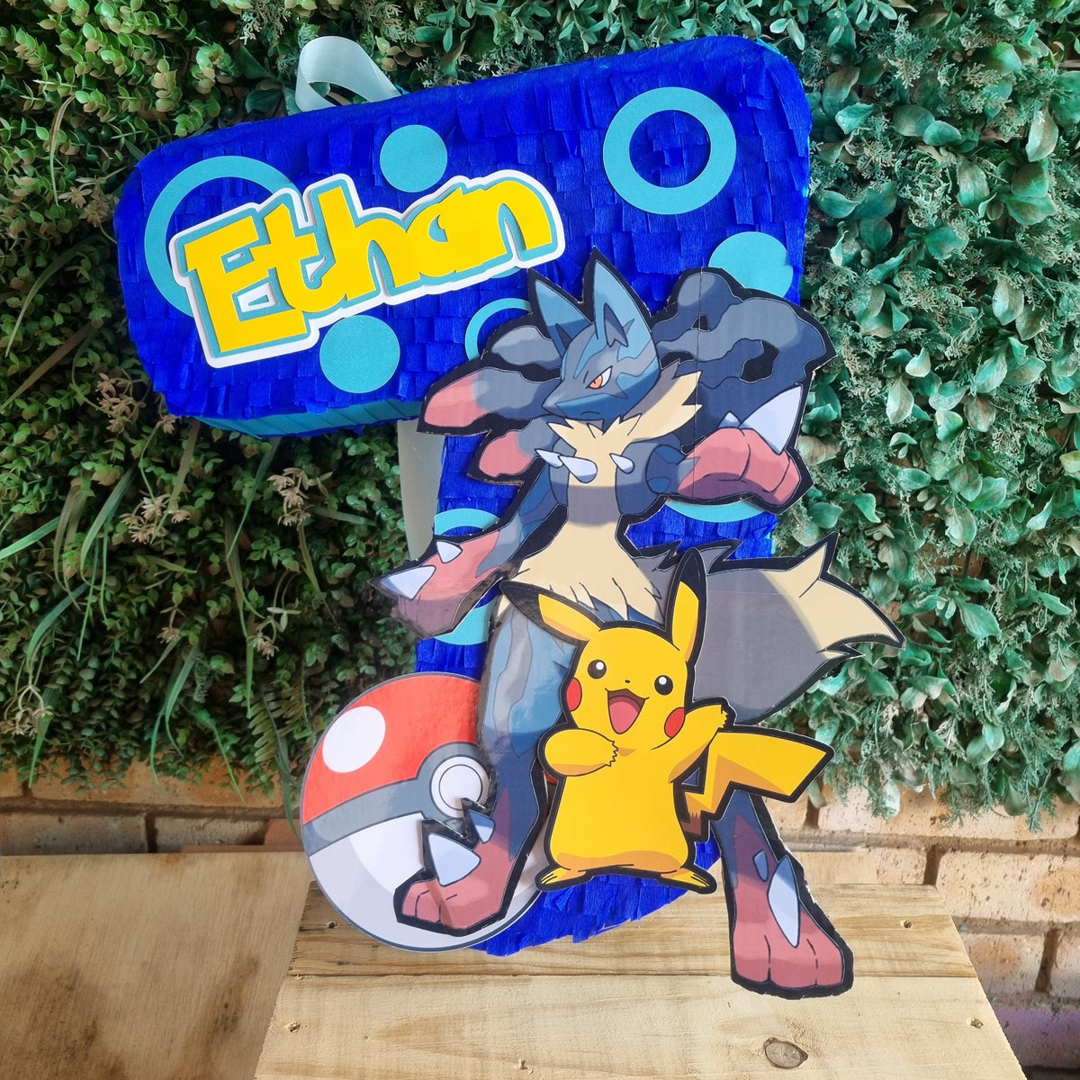 A custom Pikachu and Groudon party pinata featuring the name 'Ethan' in yellow on a blue background, displayed on a wooden surface in front of a green leaf wall.