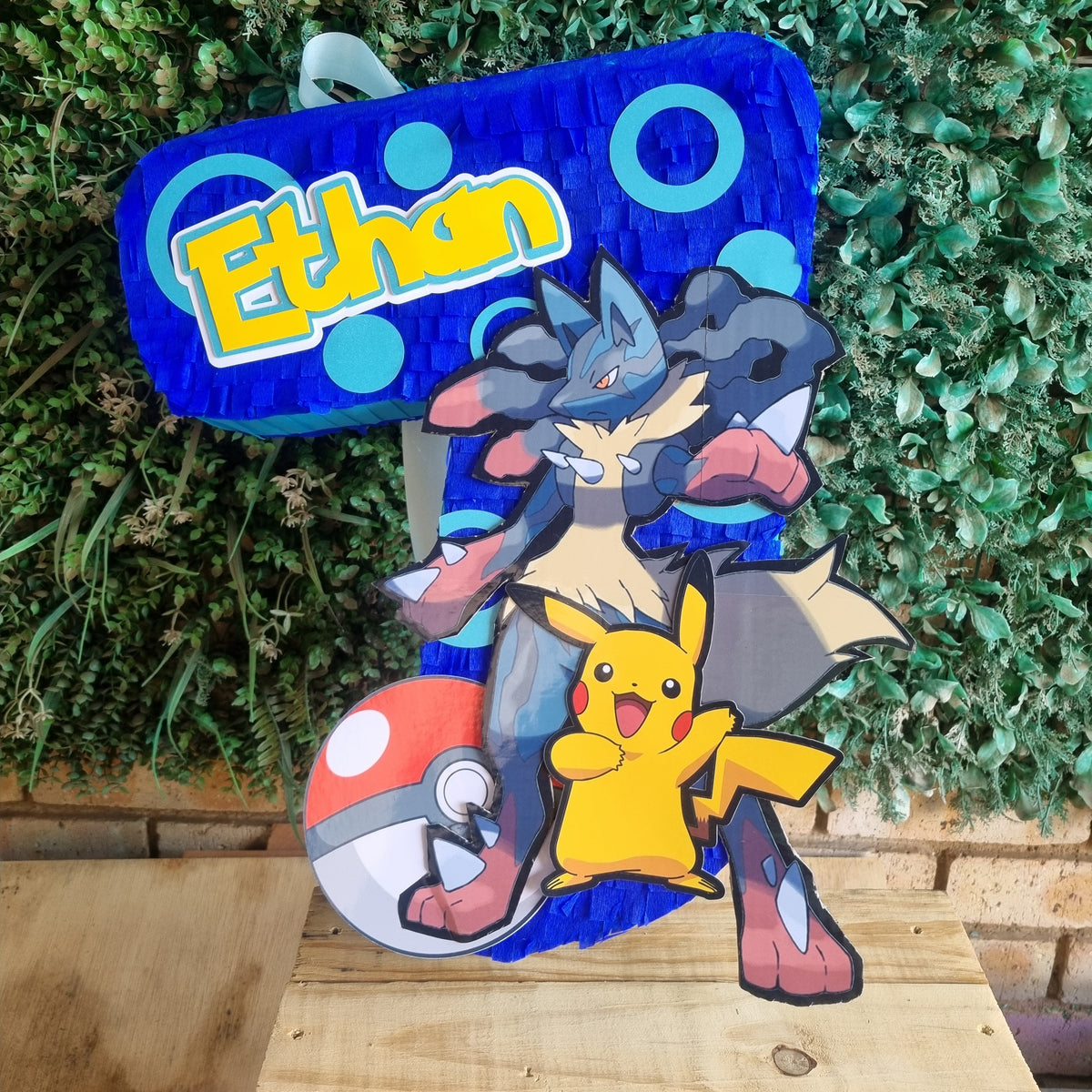 A custom Pikachu and Groudon party decoration featuring the name 'Ethan' in yellow on a blue background, displayed on a wooden surface in front of a green leaf wall.