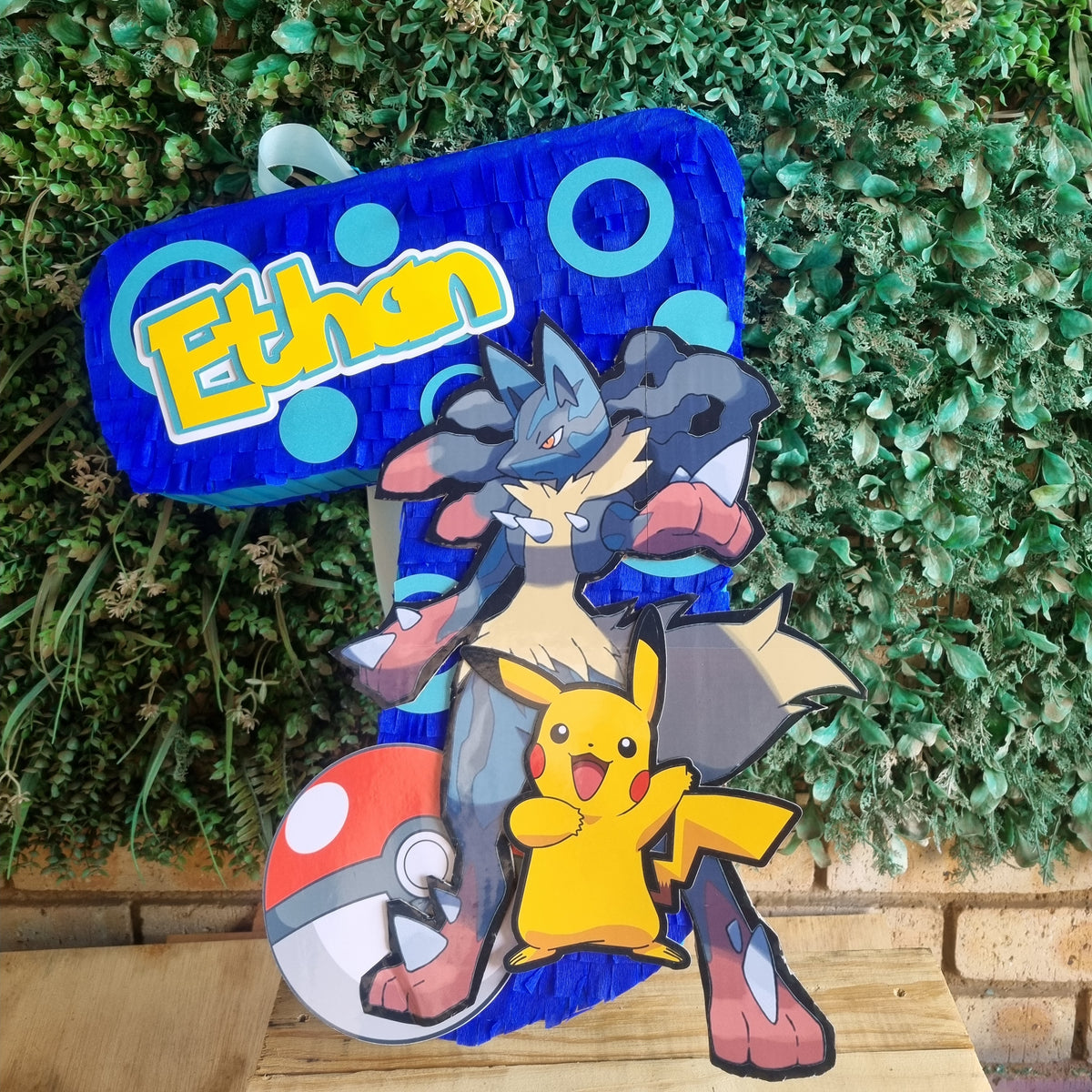 A custom cardboard party cutout featuring a blue background with the name 'Ethan' in yellow, decorated with Pokémon characters including Lucario, Pikachu, and a red-and-white ball, set against a green hedge background.