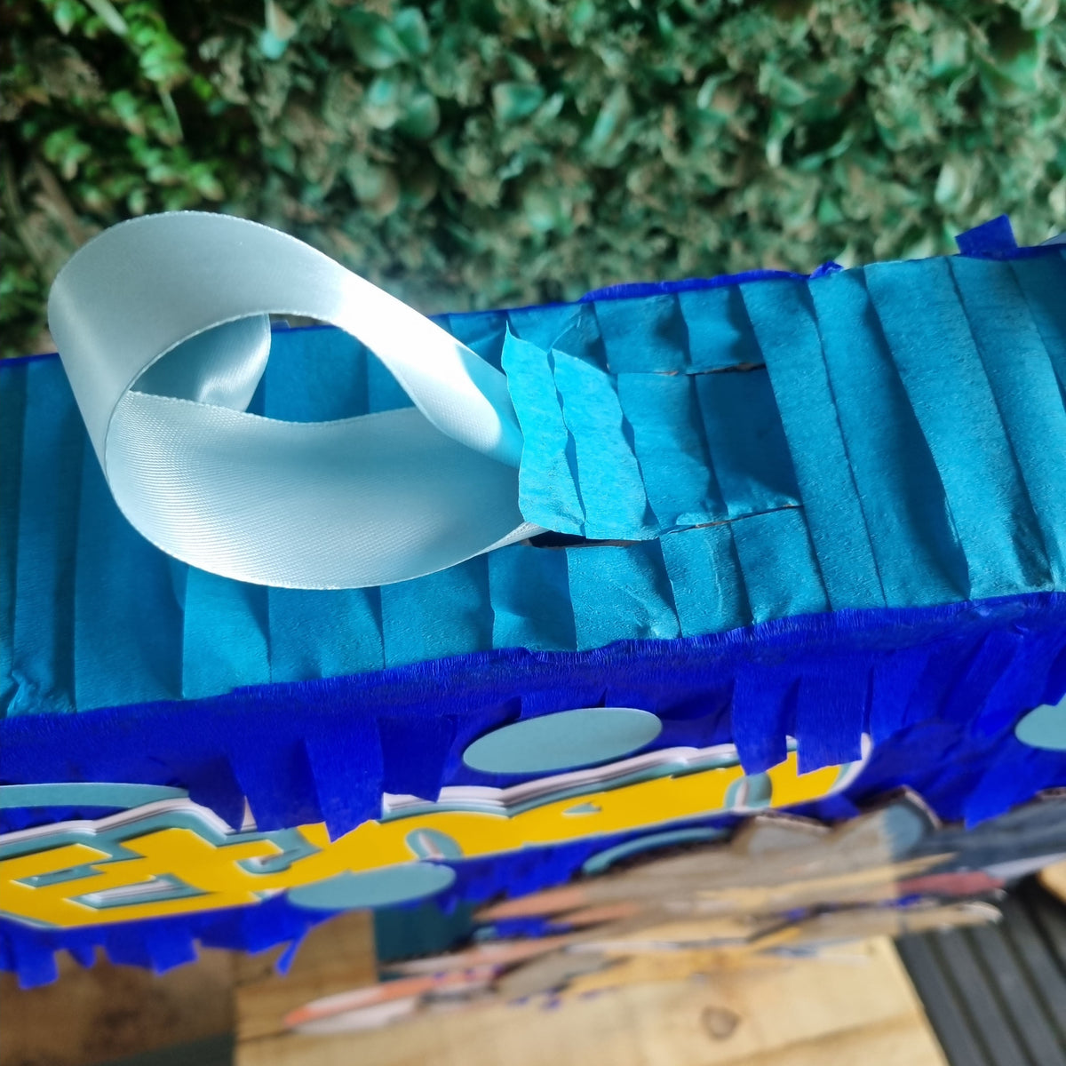 A close-up of a blue and teal fringed box or package with a white loop handle, photographed outdoors against a green hedge and wooden surface.