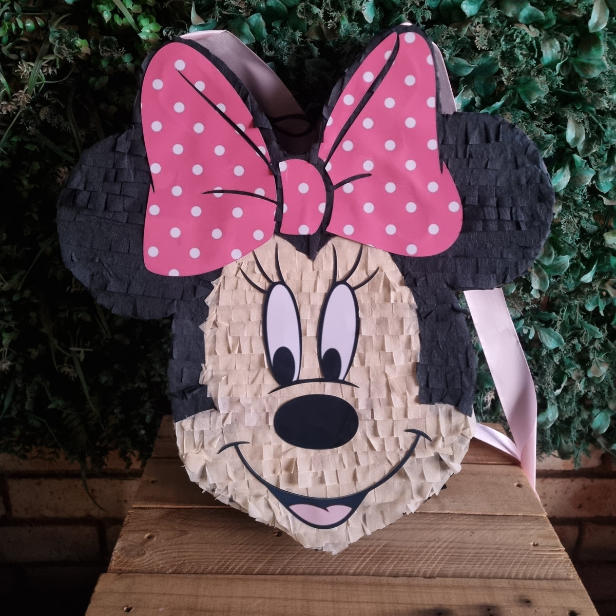 A Minnie Mouse piñata bag shaped like her face, featuring a large pink bow with white polka dots, black ears, and a textured beige paper body, displayed on a wooden surface against a green leafy background.