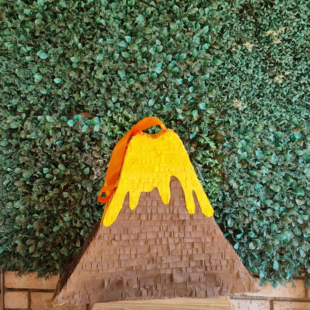 A volcano-shaped pinata with a brown textured body, a bright yellow dripping crater, and an orange peak, displayed against a green leaf wall backdrop.