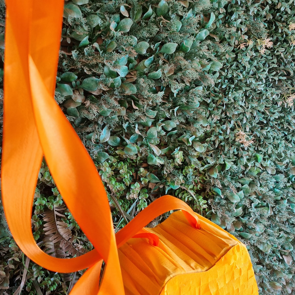 A close-up of a bright orange volcano pinata with a wide orange strap, photographed outdoors against a dense green leafy background.