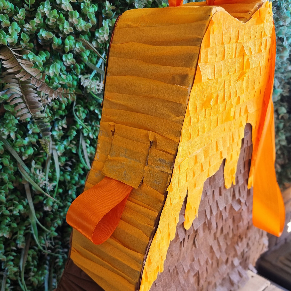 A close-up of a mountain-themed party decoration or piñata with a textured orange-yellow peak, brown mountain face, and bright orange ribbon, photographed against a green foliage backdrop.