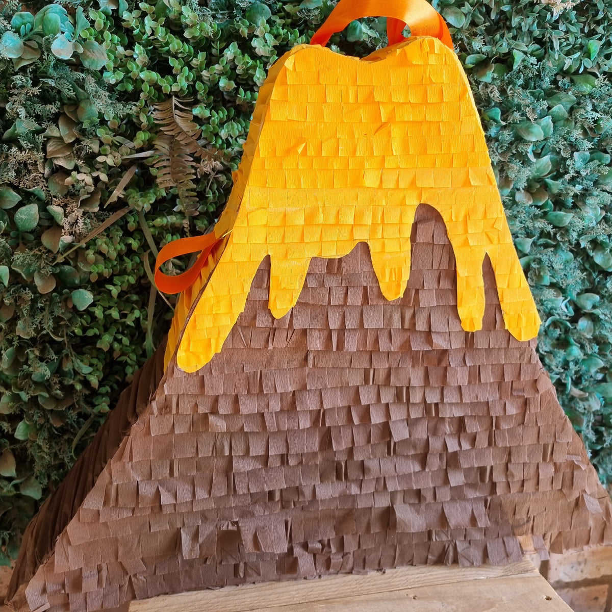 A volcano-shaped party piñata with a textured brown paper body, bright orange dripping lava, and an orange hanging loop, set against a green leafy backdrop.