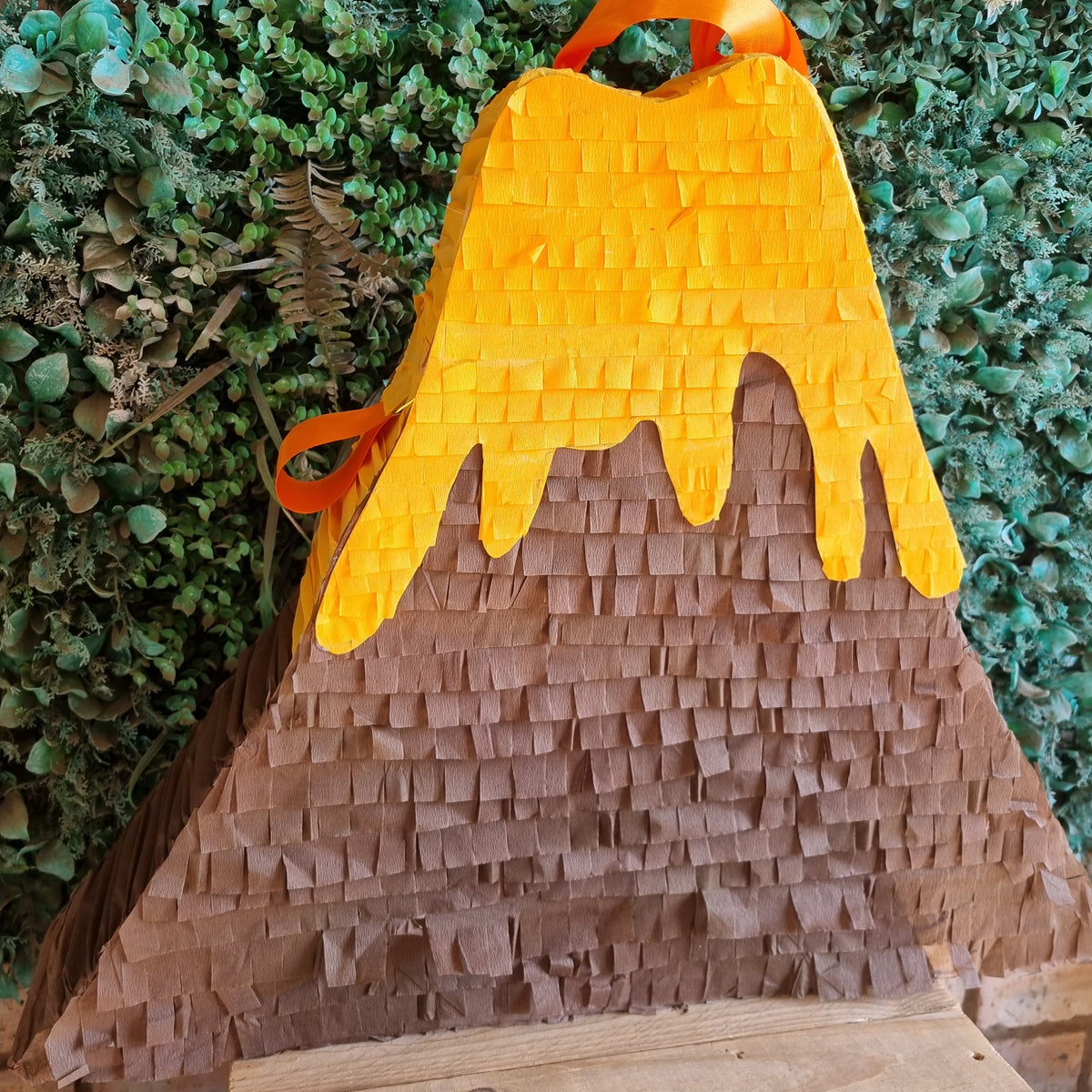 A volcano-shaped party piñata with a textured brown paper body, bright orange dripping lava, and an orange hanging loop, set against a green leafy backdrop.