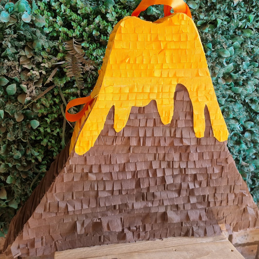A volcano-shaped party piñata with a textured brown paper body, bright orange dripping lava, and an orange hanging loop, set against a green leafy backdrop.