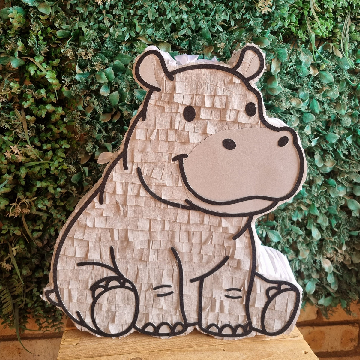A hippo-shaped pinata made of textured paper, displayed on a wooden surface in front of a green leafy wall.