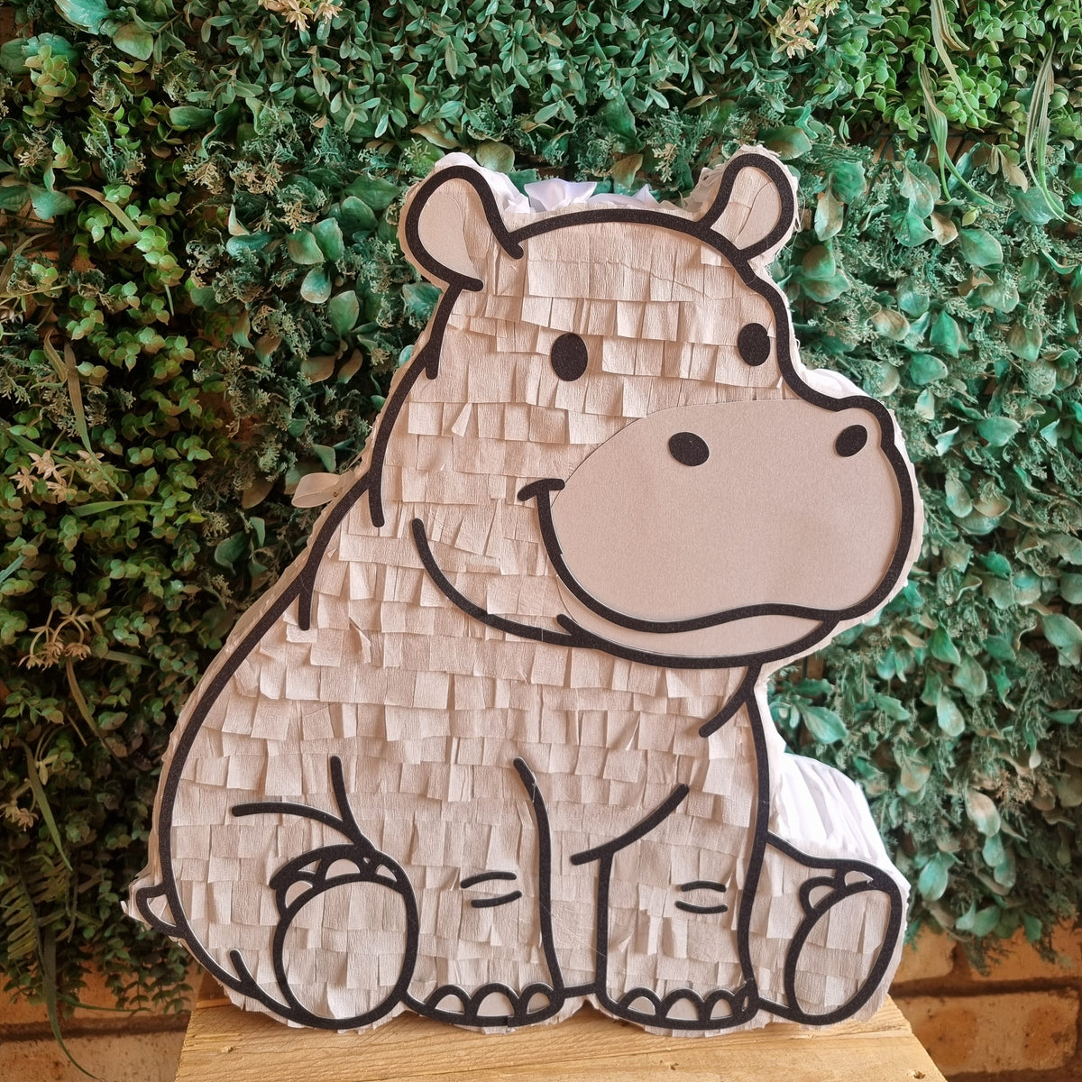 A hippo-shaped pinata made of textured paper, displayed on a wooden surface in front of a green leafy wall.