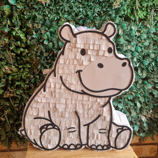 A hippo-shaped pinata made of textured paper, displayed on a wooden surface in front of a green leafy wall.