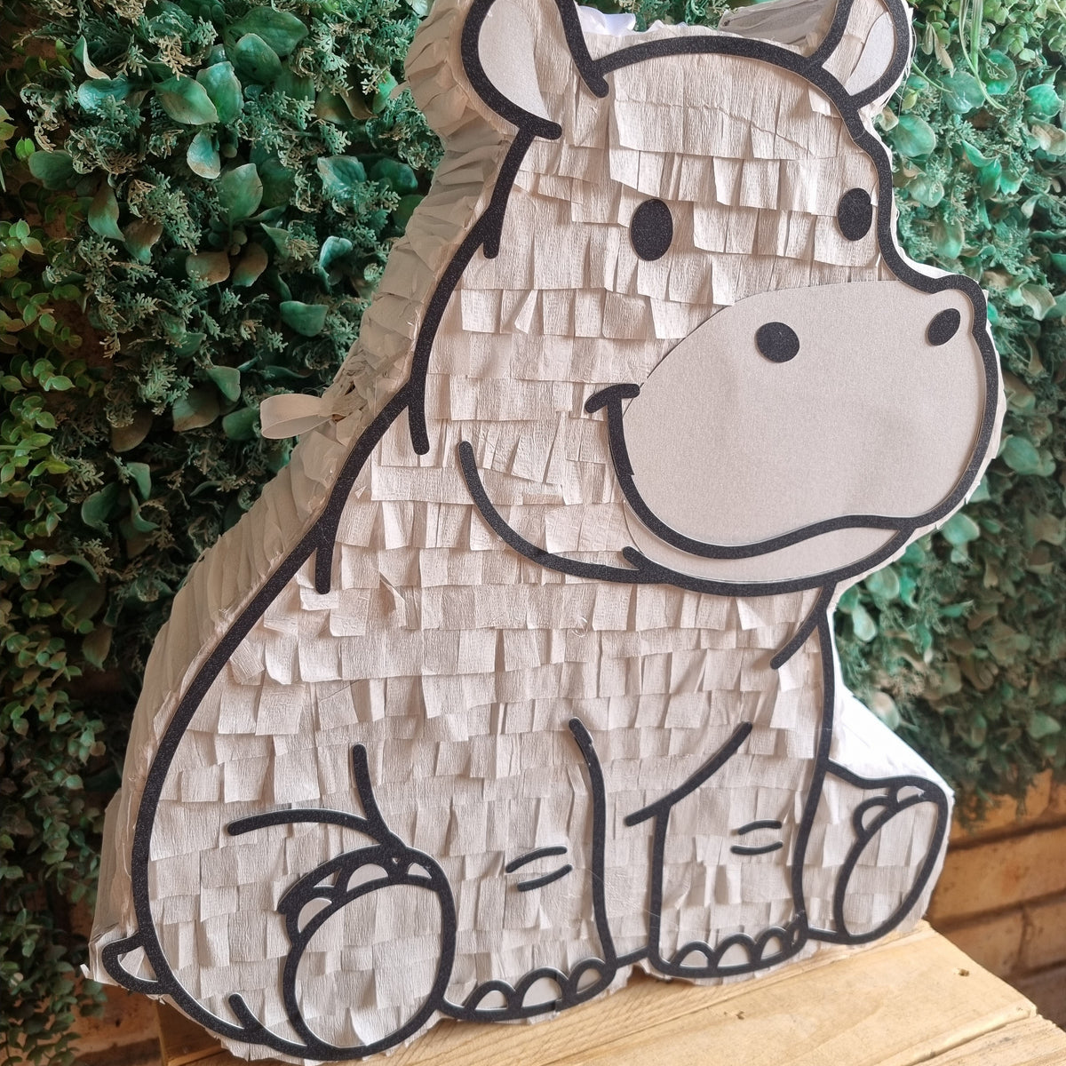 A hippo-shaped paper pinata party decoration with a textured white and beige paper body, outlined in black, displayed on a wooden surface in front of a green leafy wall.