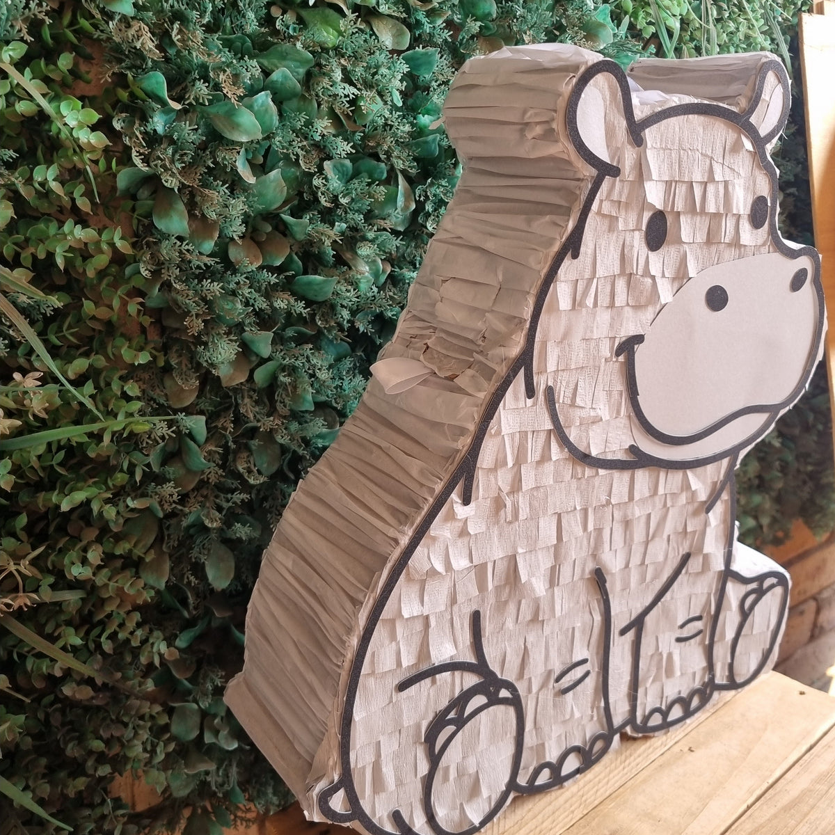 A hippo-shaped decorative sign or pinata made from textured cardboard or paper, displayed on a wooden crate in front of a green leafy wall.