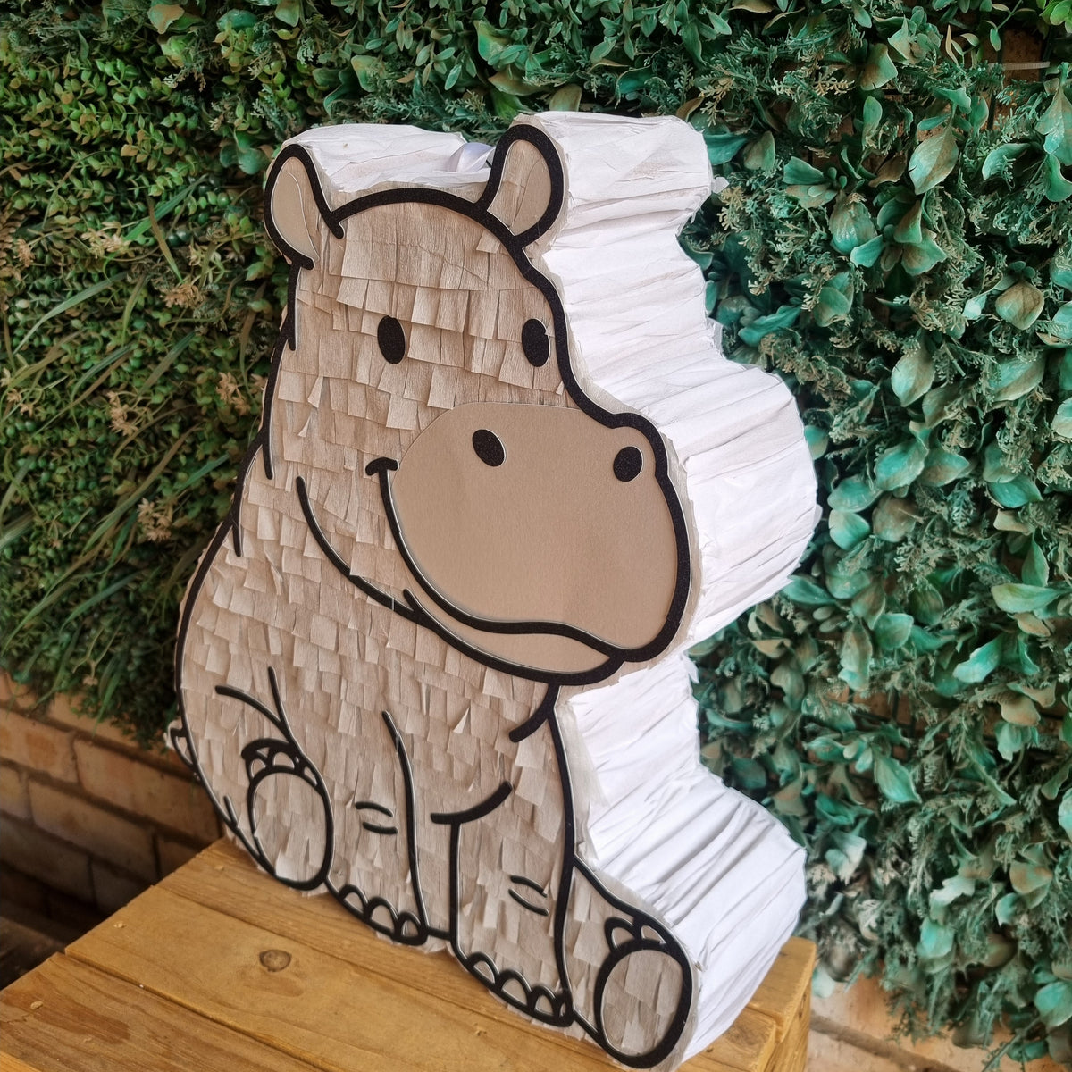 A hippo-shaped party piñata made of stacked white paper with a tan face and black outlines, displayed on a wooden stand in front of a green leafy wall.