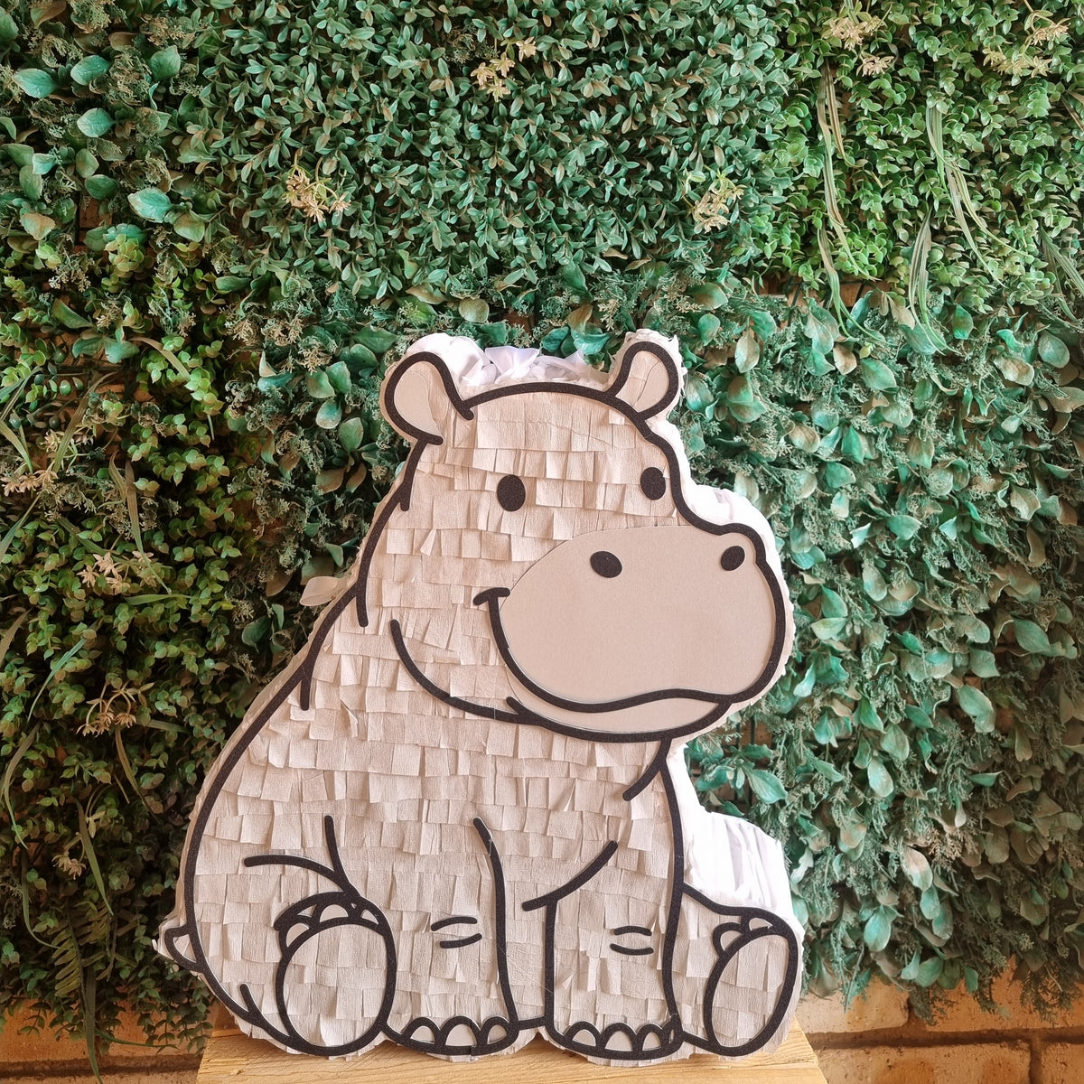 A cute cartoon hippo piñata (party hat) made of textured paper, set against a green leafy wall background.