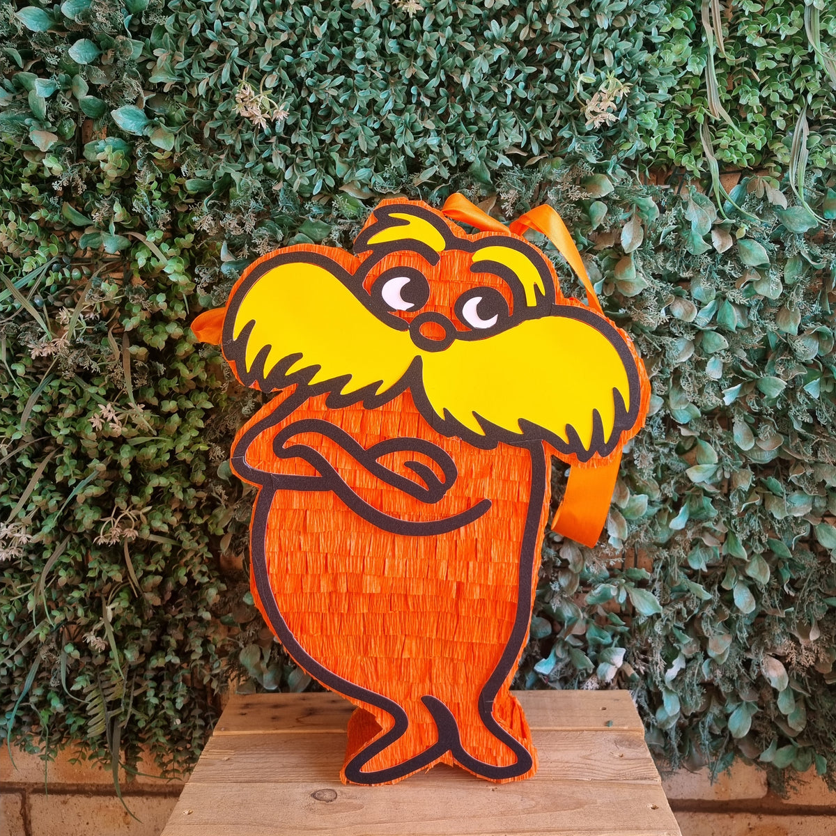 A Dr. Seuss-themed piñata figure of the Lorax standing on a wooden platform in front of a green leafy wall.