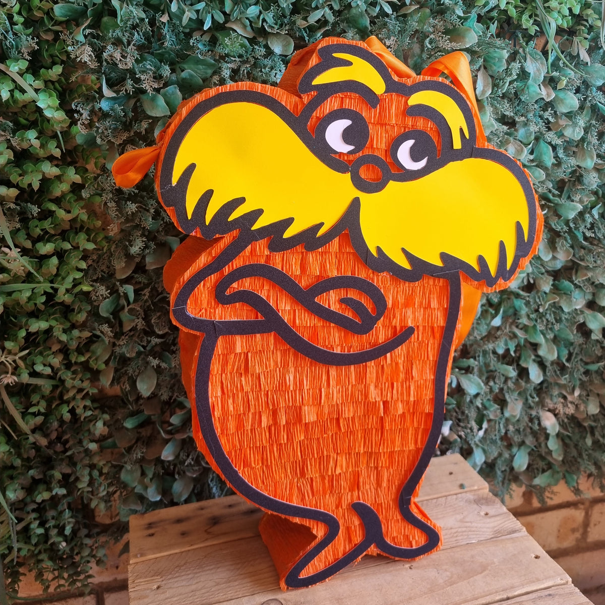 An orange and yellow LORAX character piñata (decorative target) with a textured paper finish, standing on a wooden surface against a green leafy background.