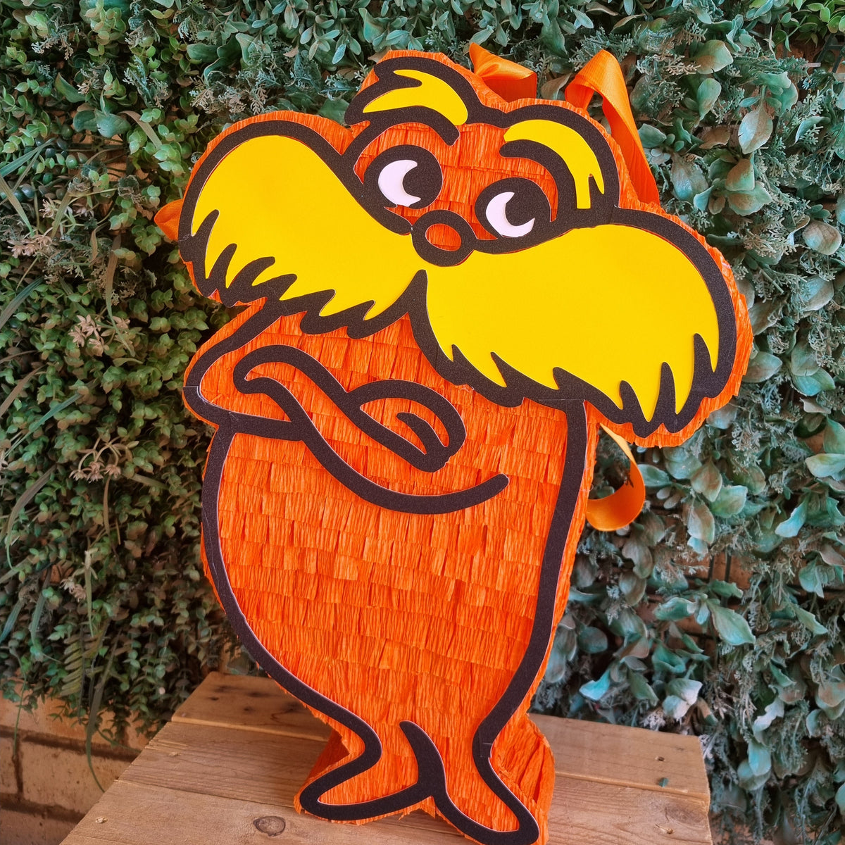 An orange and yellow Dr. Seuss-themed pinata shaped like the Lorax standing on a wooden surface in front of a green hedge background.