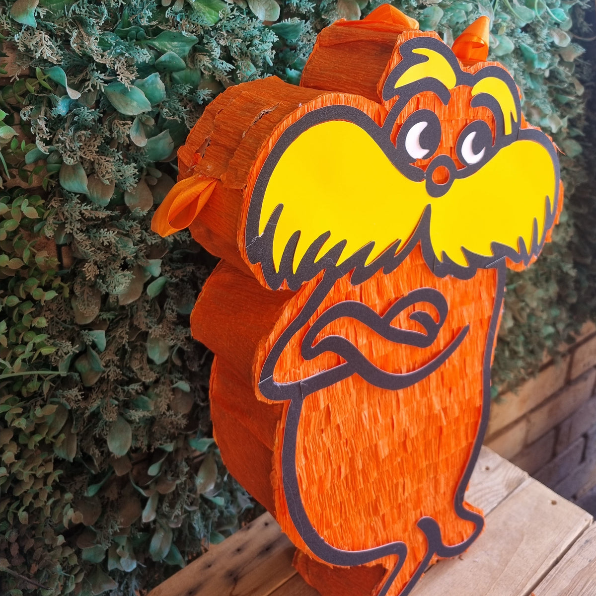 A LORAX figurine painted with a bright orange body and a large yellow beard, standing on a wooden surface against a green leafy wall background.