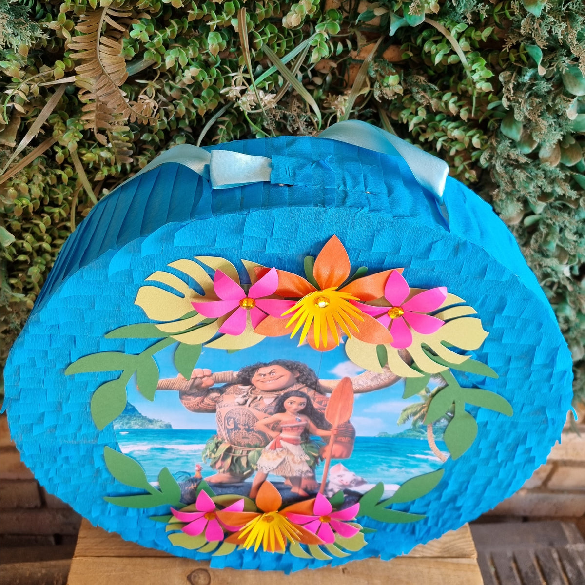 A round blue pinata featuring a Polynesian warrior illustration surrounded by colorful paper flowers and green leaves, set against a green foliage background.