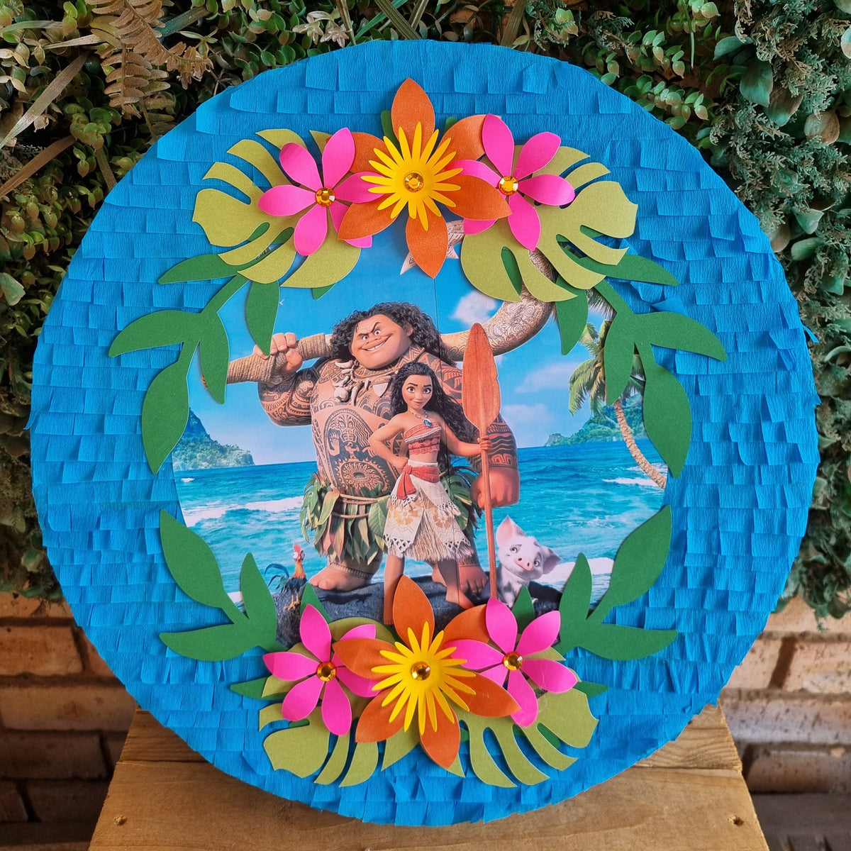 A round Moana-themed pinata displayed outdoors on a wooden stand, featuring colorful paper flowers, green leaves, and characters from the movie against a bright blue background.