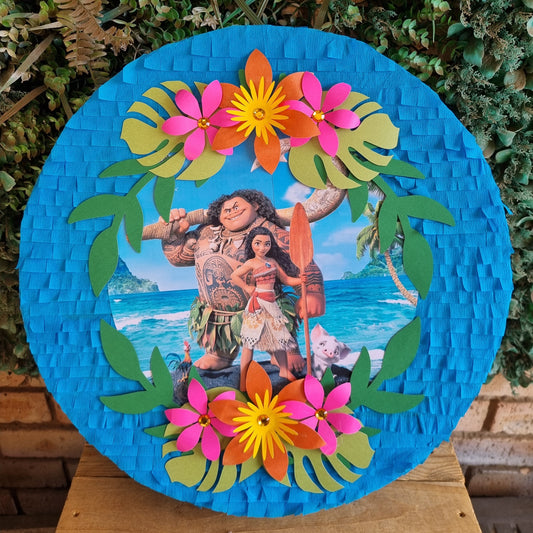 A round Moana-themed pinata displayed outdoors on a wooden stand, featuring colorful paper flowers, green leaves, and characters from the movie against a bright blue background.