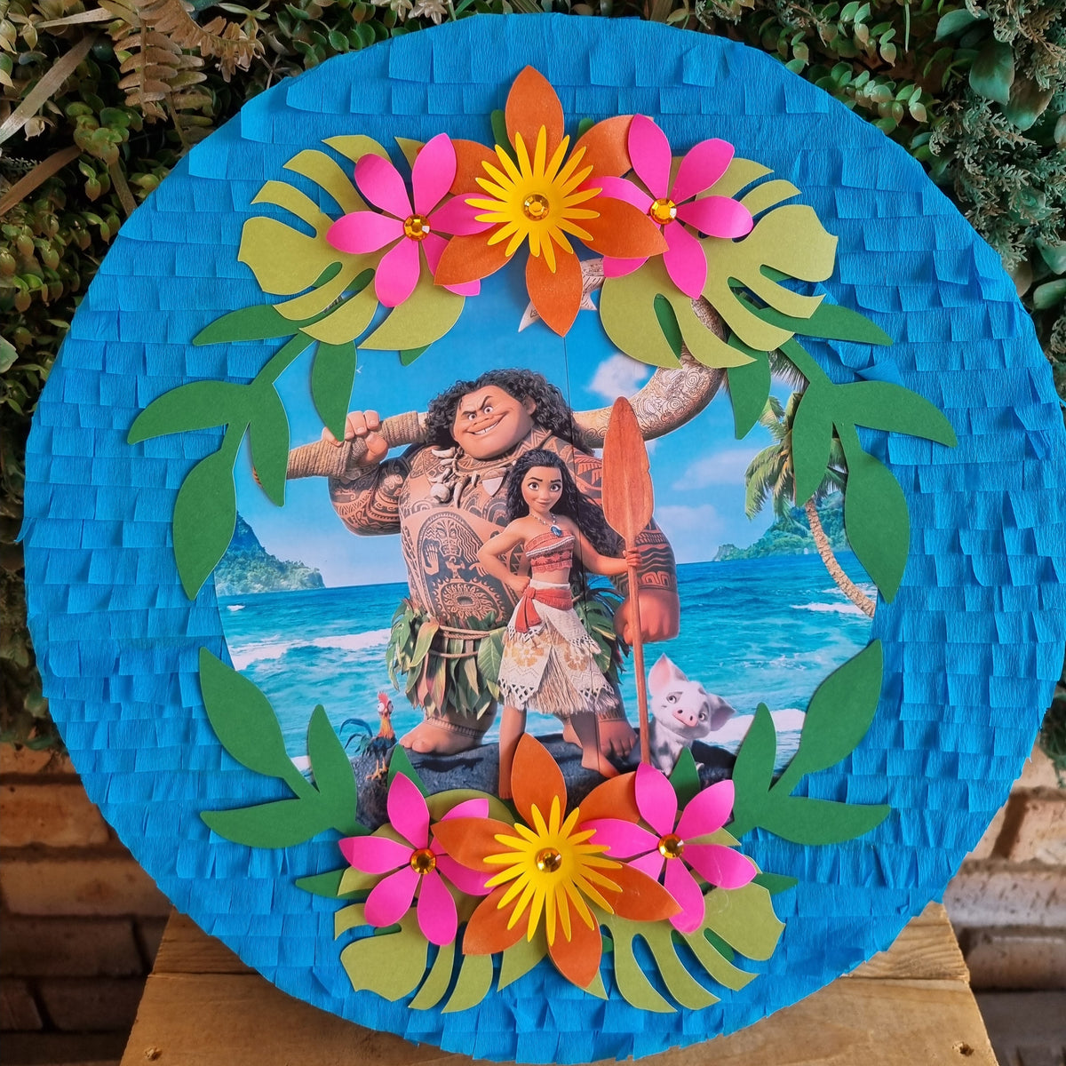 A circular Moana-themed pinata displayed outdoors on a wooden surface, featuring a blue woven border, colorful paper flowers, green leaves, and a central photo of Moana and Maui on a beach.