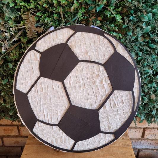 A round soccer ball themed pinata displayed on a wooden surface in front of a green leafy wall.