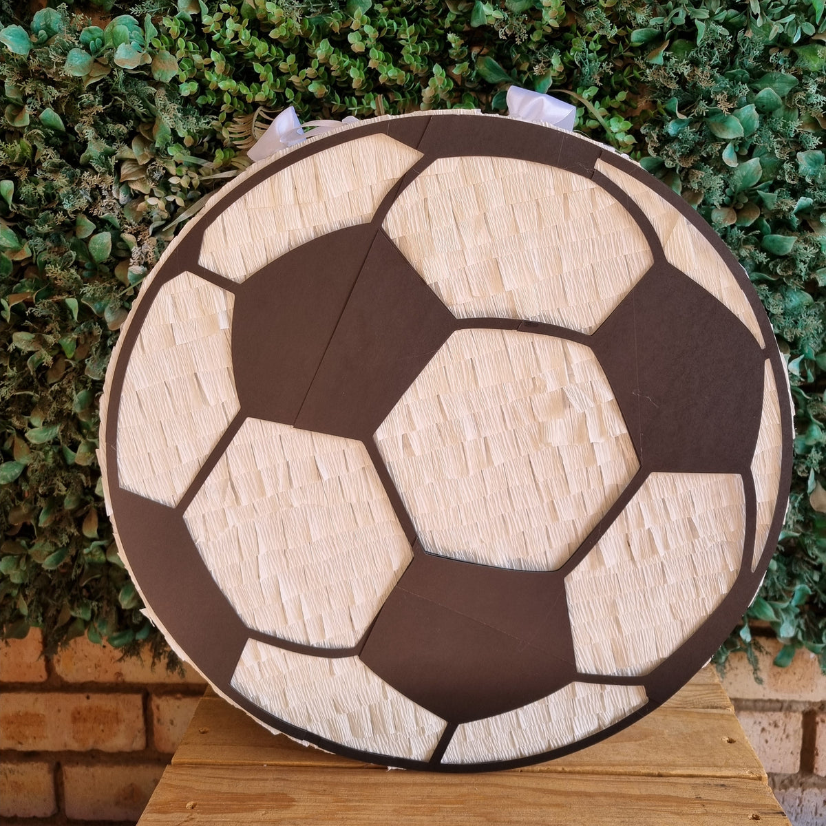 A round soccer ball themed party piñata decoration displayed on a wooden table in front of a green leafy hedge wall.