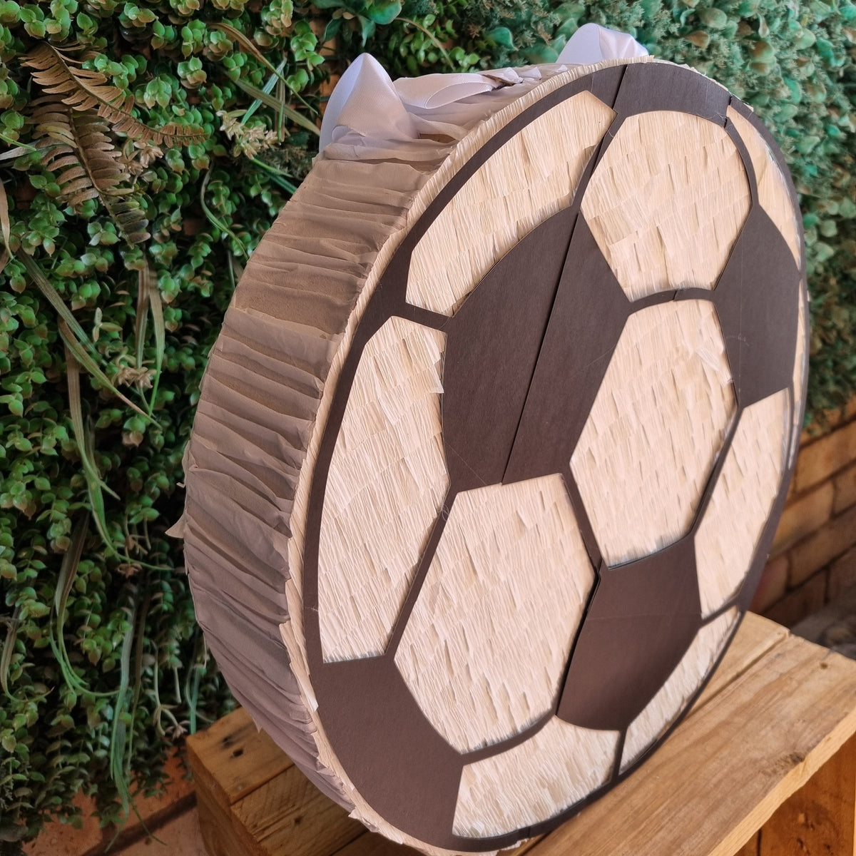 A round, soccer-ball-shaped party piñata decoration with a dark brown and light beige pattern, wrapped in clear plastic on the sides, displayed on a wooden stand against a green leafy background.