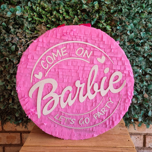 A round pink Barbie-themed party piñata sign with white lettering that reads 'COME ON Barbie LET'S GO PARTY,' displayed on a wooden surface against a green leafy hedge background.