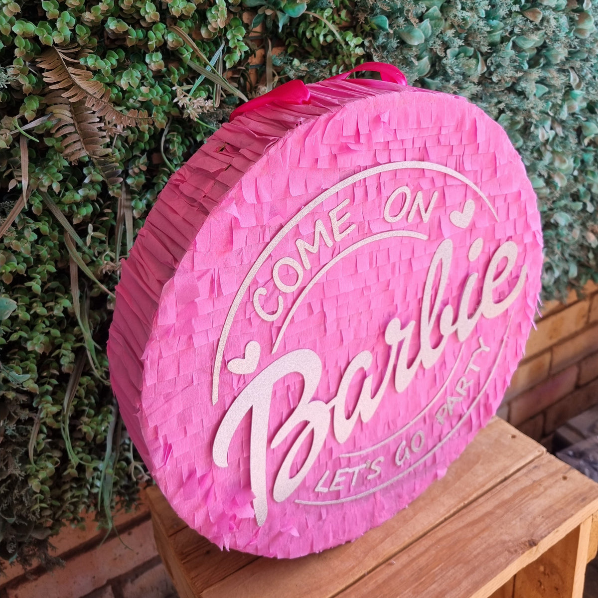 A pink fringed Barbie-themed party decoration sign on a wooden crate in front of a green hedge wall. The sign features the text 'COME ON Barbie LETS GO FIRST' in white.