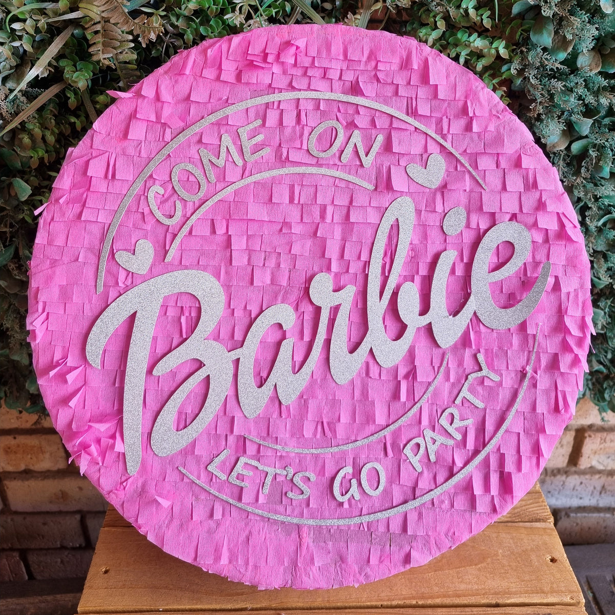 A round pink fringed pinata with 'COME ON Barbie LET'S GO PARTY' in white lettering, displayed on a wooden surface in front of a green leafy background.