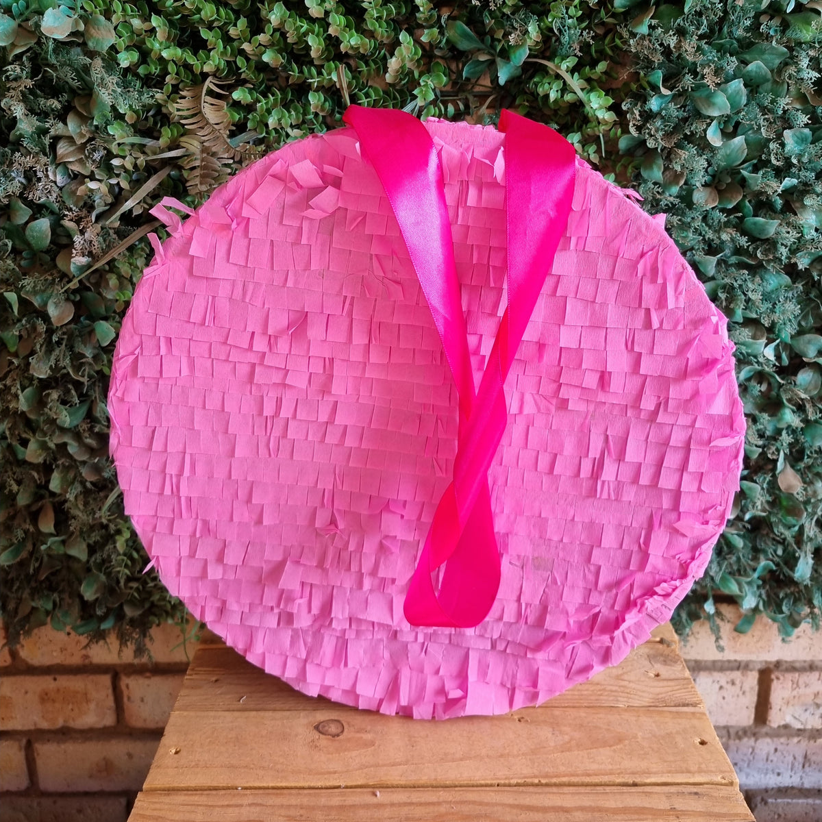 A round pink paper piñata with a bright pink ribbon bow, displayed on a wooden table in front of a green leafy wall.