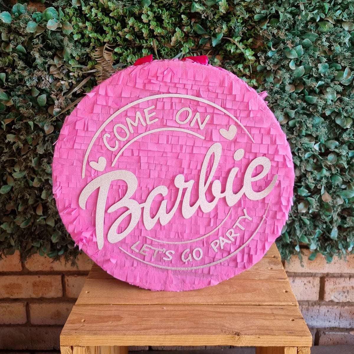 A round pink Barbie-themed party piñata sign with white lettering that reads 'COME ON Barbie LET'S GO PARTY', displayed on a wooden stand in front of a green hedge background.