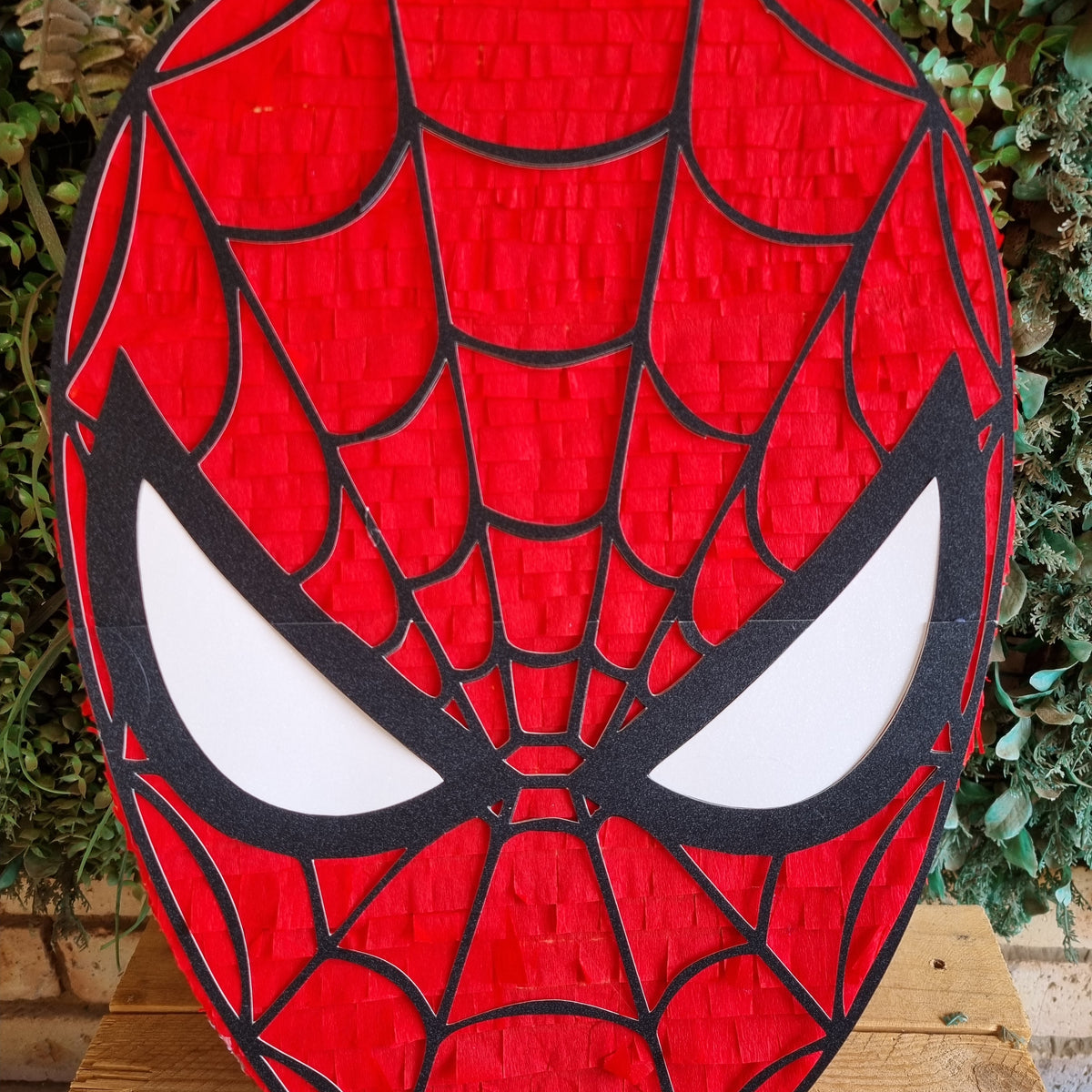 A red Spider-Man themed pinata shaped like his mask, featuring black web lines and white eyes, displayed on a wooden surface with green foliage in the background.
