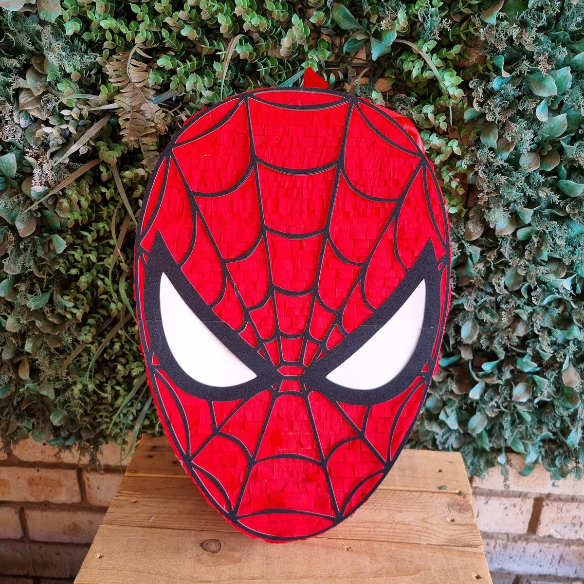 A red and black Spider-Man mask-shaped decorative sign or plaque with white eyes, displayed on a wooden surface in front of a green leafy wall.
