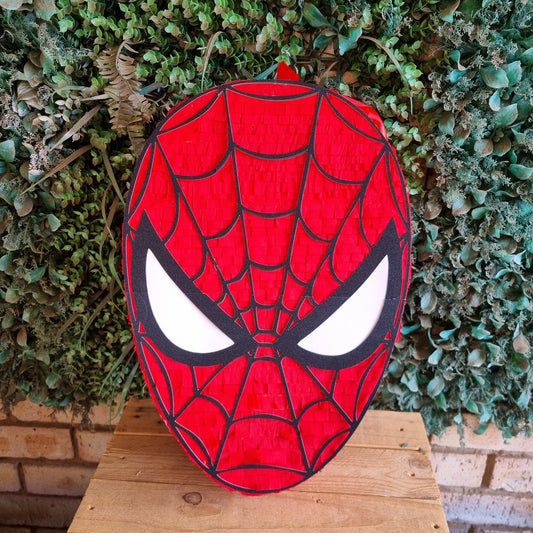 A red and black Spider-Man mask-shaped decorative sign or plaque with white eyes, displayed on a wooden surface in front of a green leafy wall.