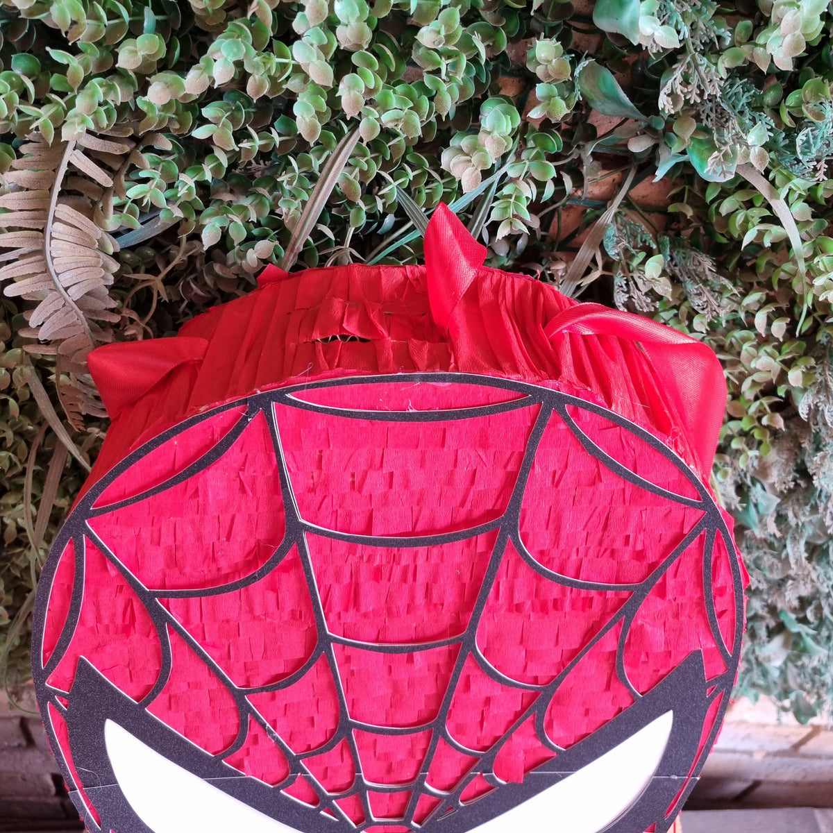 A Spider-Man themed party decoration, likely a piñata or backdrop panel, featuring a red and black spider web design with white eyes, set against a green leafy background.