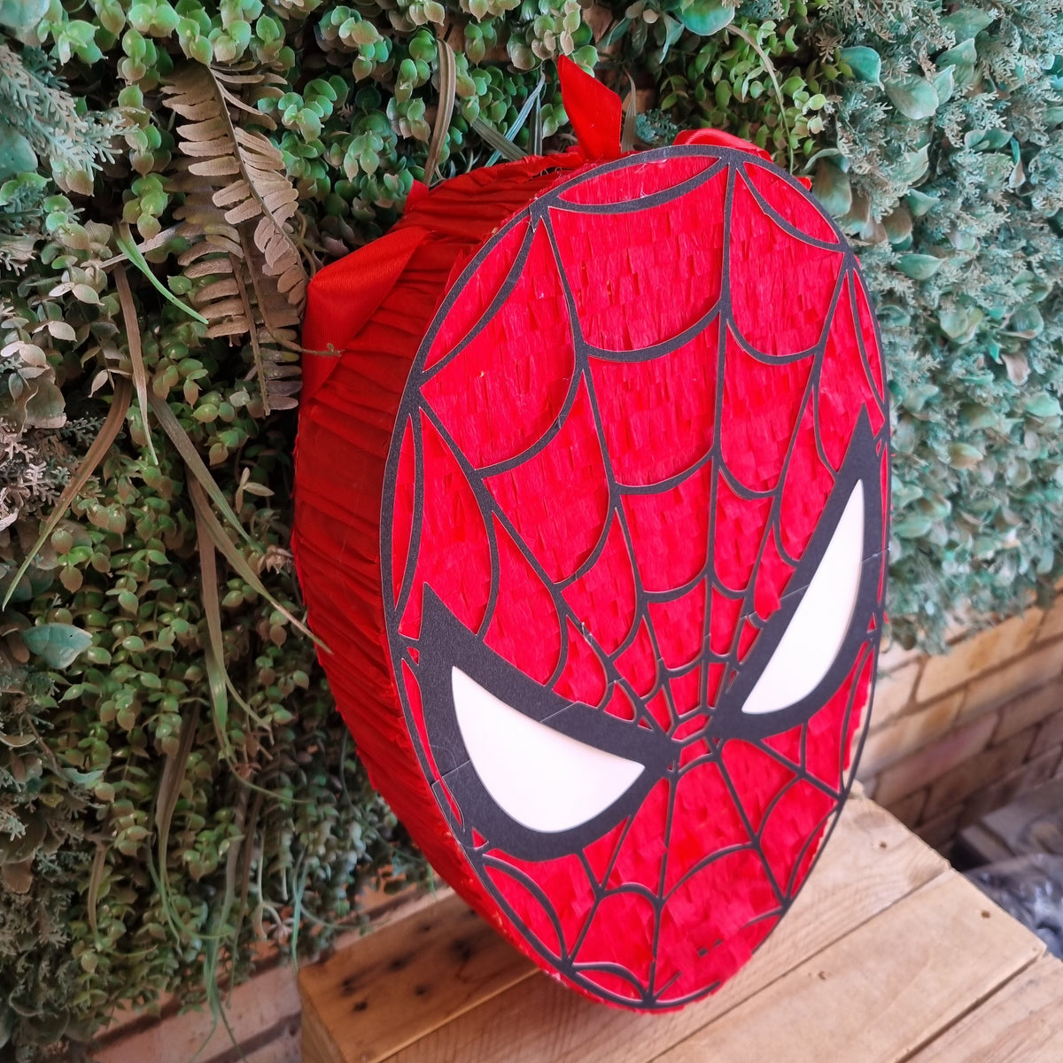 A red Spider-Man themed round lantern or decorative light shaped like his mask, leaning on a wooden crate in front of a green leafy wall.
